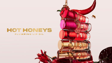 HOT HONEYS PLUMPING LIP OIL — six stacked Hot Honeys Plumping Lip Oil tubes in burgundy, pink, coral, and nude shades with a chili pepper and honey drizzle.