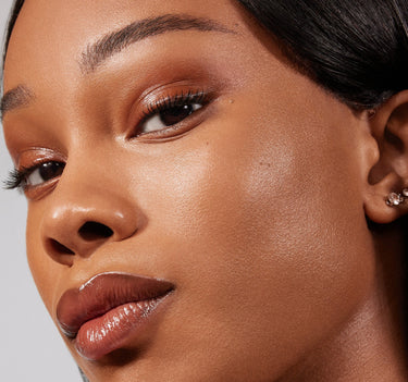{Rich 24W} Close-up of Lightform Extended Hydration Foundation on deep skin tone, showcasing a luminous, glass-skin finish with warm bronze eye and glossy nude lip.