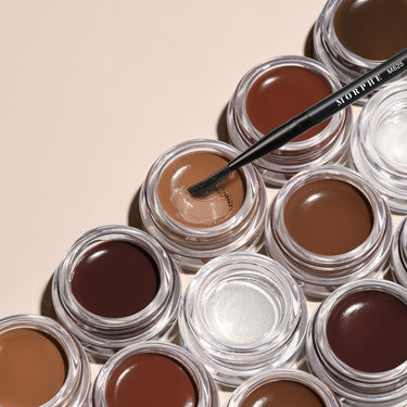 Stylized group of Morphe Supreme Brow Sculpting and Shaping Wax jars in shades from blonde to espresso, with a MORPHE M625 brush.