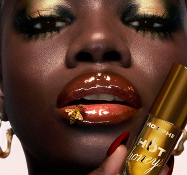 {Royal Sting} Close-up of deep bronzed-gloss plumping lip oil on dark skin with a gold bee detail on lips, holding the gold Morphe Hot Honeys tube.